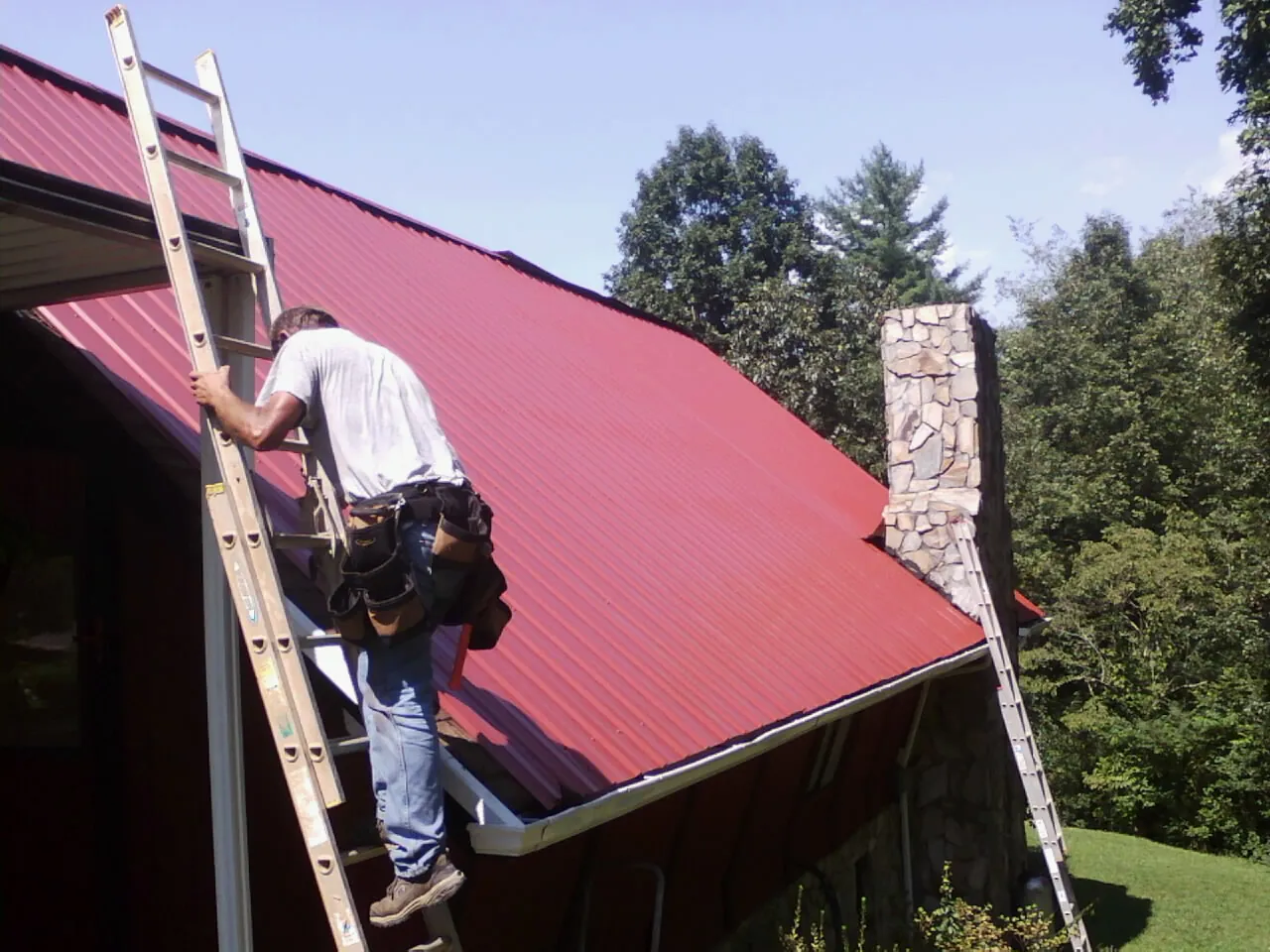 Roofer on a ladder beside a red metal roof and stone chimney — Soffit & Fascia Repair by Roofing Brothers in Lunenburg