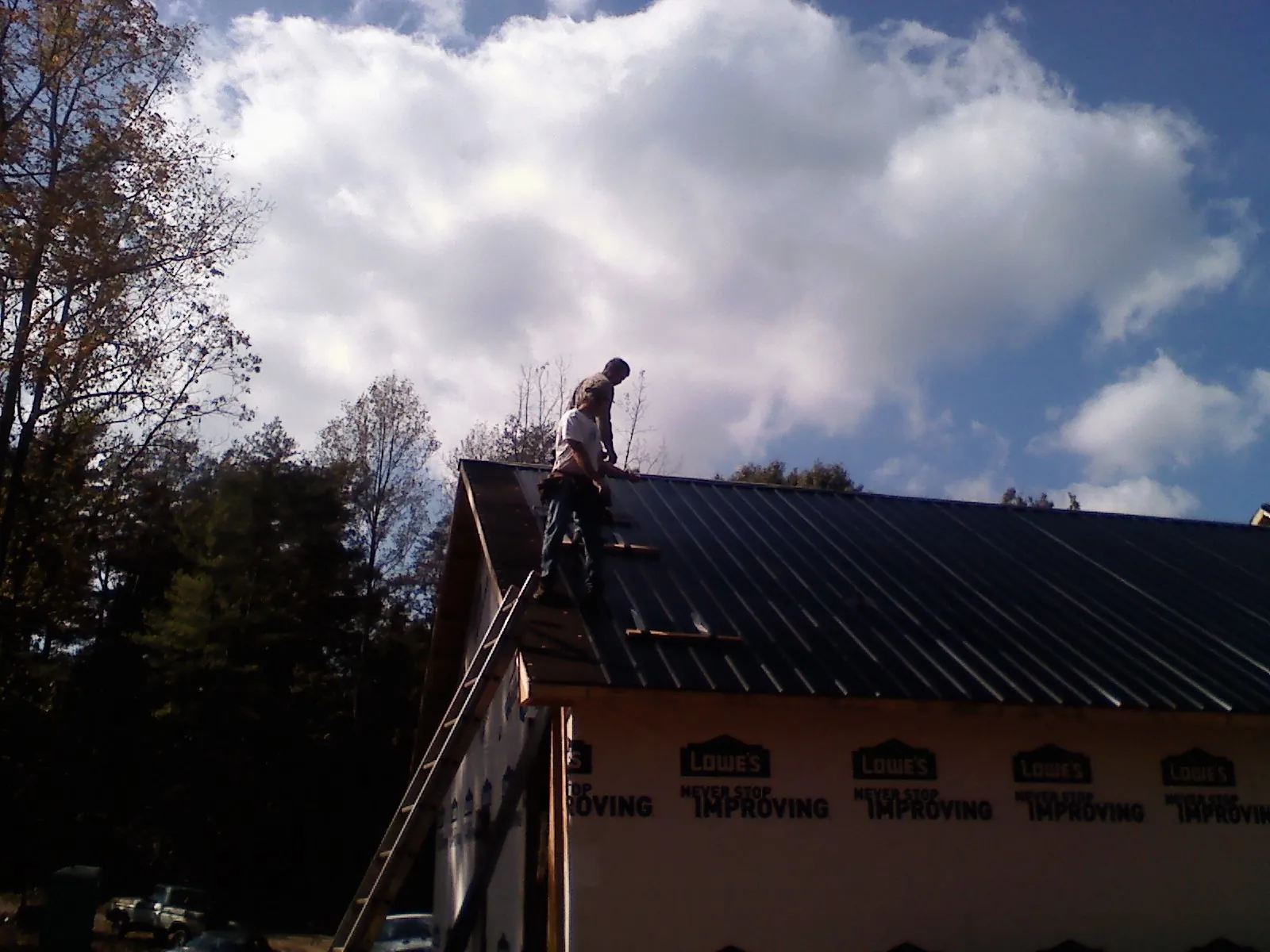 Roofer fastening dark metal panels high against a cloudy sky, from a Roof Inspection job in Lunenburg